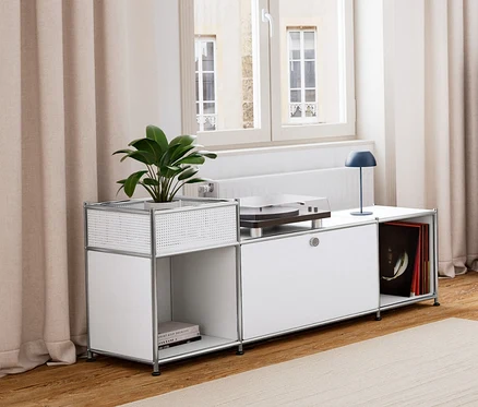 Salon z drewnianą podłogą i jasnym dywanem. Po lewej biała metalowa szafka typu lowboard „CN3” z klapą, na niej granatowa akumulatorowa lampa stołowa LED. Obok granatowa metalowa komoda „CN3” z 3 klapami. Na środku stolik z tacą i funkcją siedziska oraz fotel. W tle szara metalowa szafka typu sideboard „CN3” z 3 klapami.