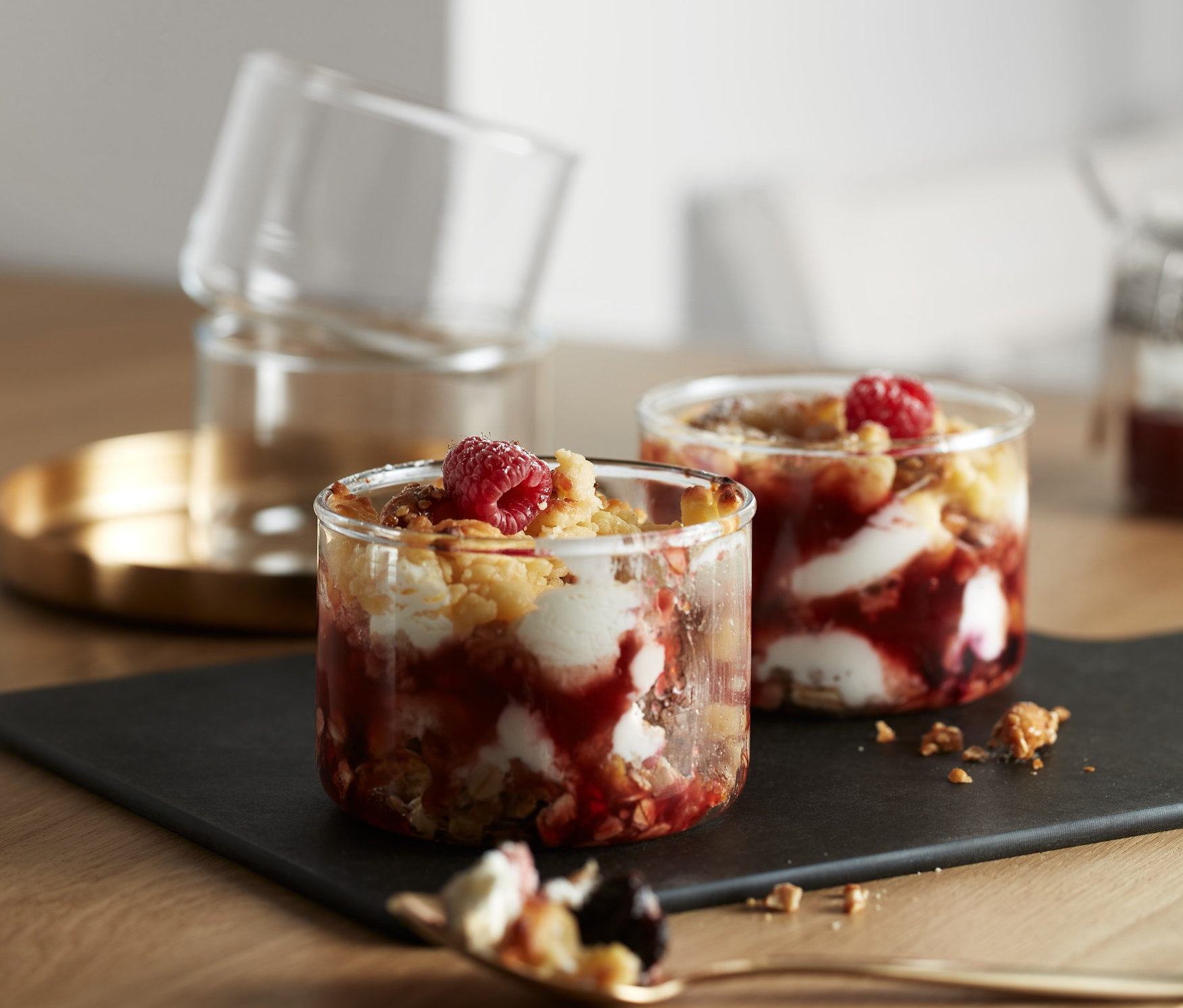 Na stole stoją 4 sztuki foremek do porcjowania i pieczenia z deserem owocowym. Dwie foremki stoją na czarnej podkładce, a dwie pozostałe są ułożone jedna na drugiej na złotej tacy.