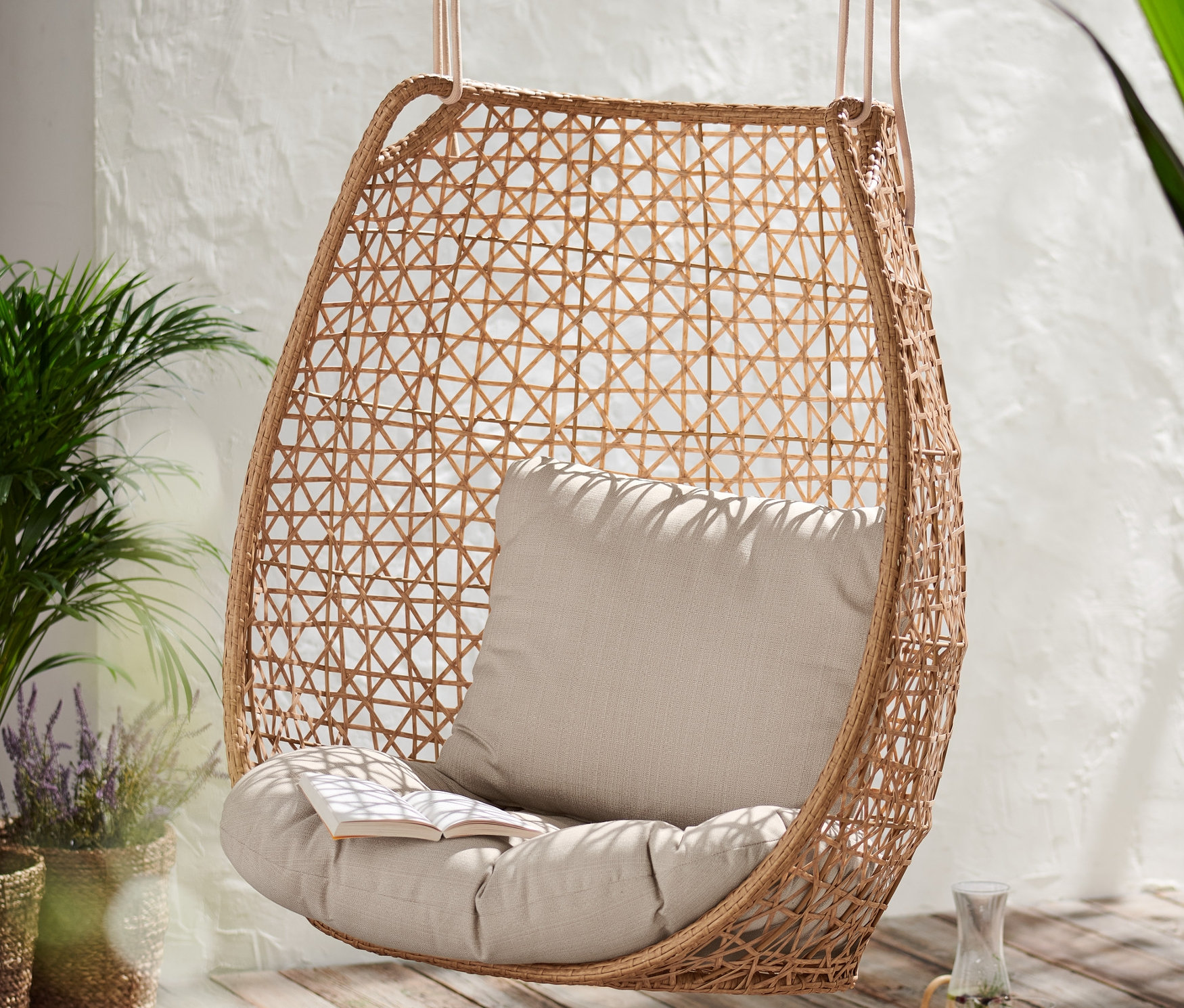 Fotel wiszący »boho« z beżową poduszką i książką na drewnianej podłodze.