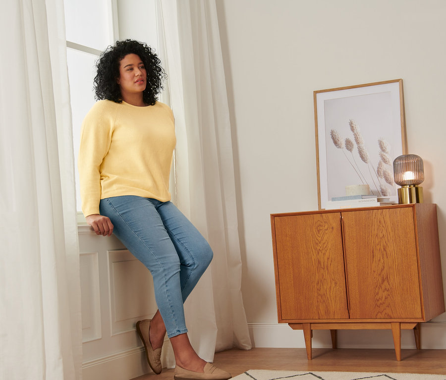 Kobieta siedzi na parapecie w żółtym swetrze z dzianiny, tregginsach i półbutach ze skóry welurowej.