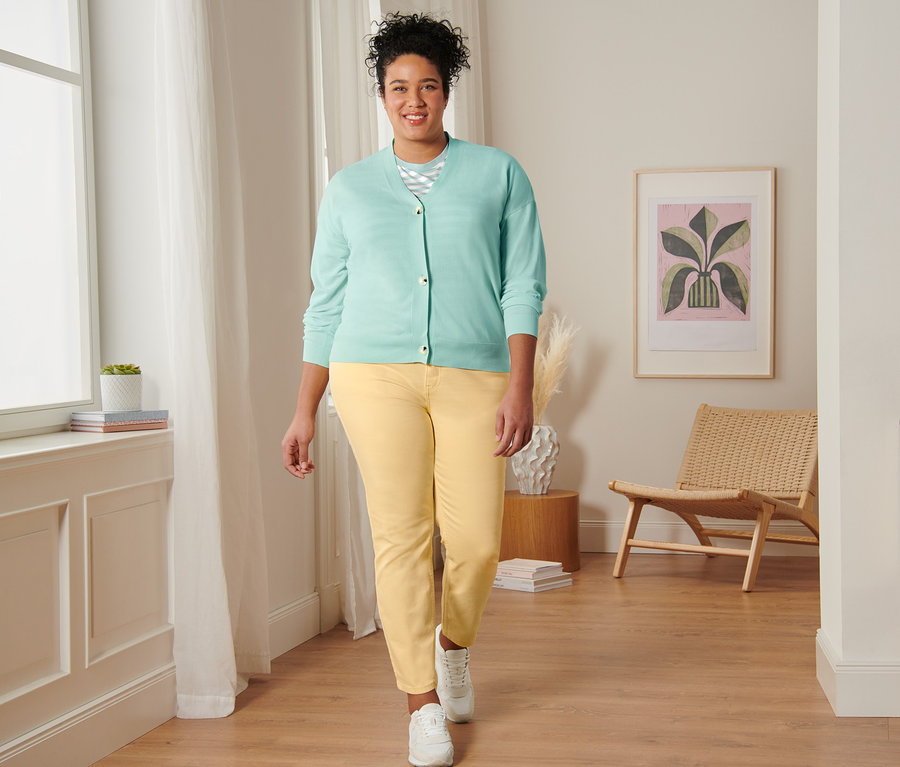 Kobieta idzie, ubrana w miętową koszulkę w paski, kardigan z wycięciem w serek, żółte spodnie dżinsowe o kroju slim fit i buty typu sneakers ze skóry welurowej.