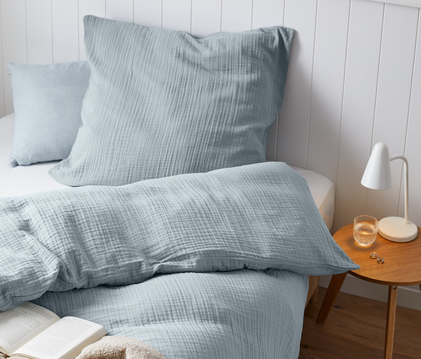 Jasnoniebieska pościel muślinowa na łóżku z otwartą książką i beżowym kocem.