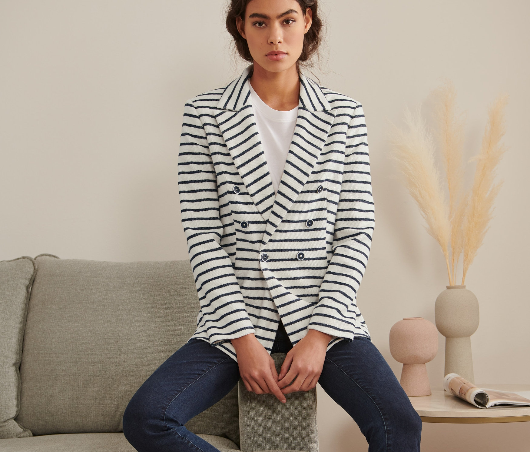 Kobieta siedzi na szarej sofie i ma na sobie żakiet z dzianiny dresowej w paski.