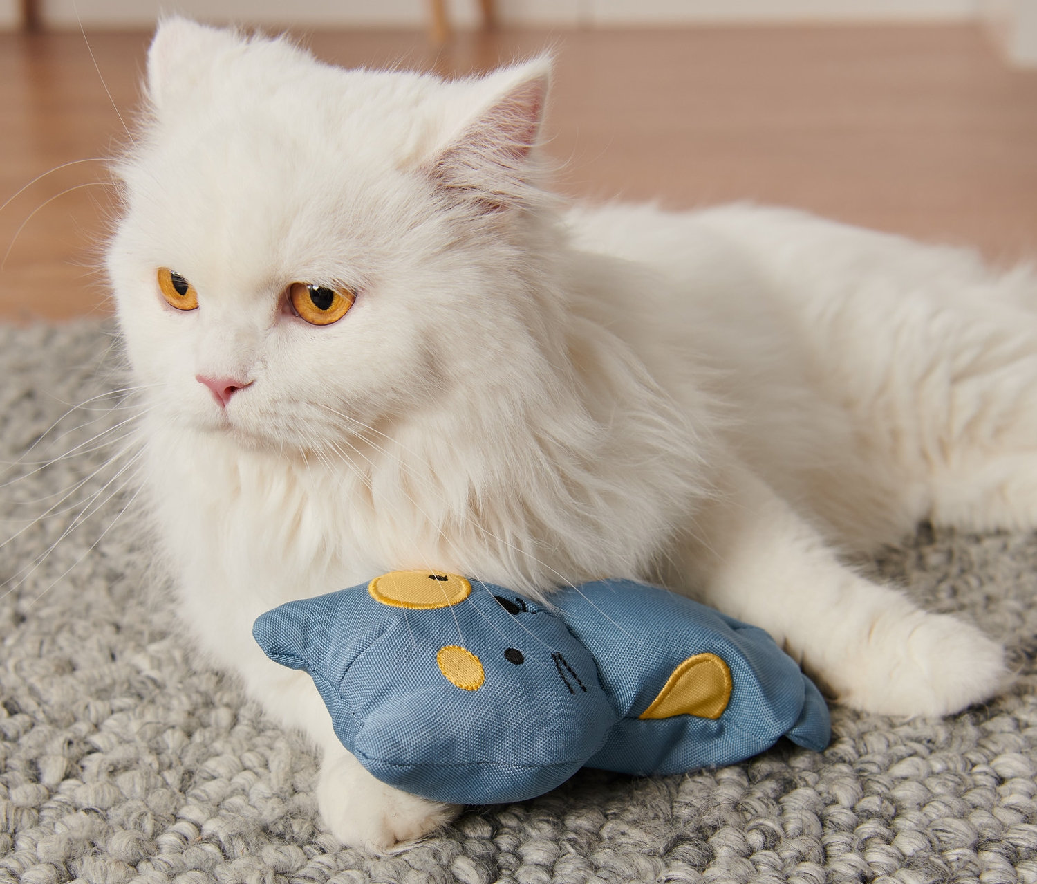 Biały kot leży na szarym dywanie i trzyma łapą niebieską zabawkę dla kota z kocimiętką.