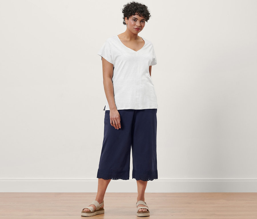 Kobieta pozuje w kremowobiałej bluzce z mieszanki materiałowej z zawartością lnu, granatowych kulotach z haftem i skórzanych sandałach na platformie.