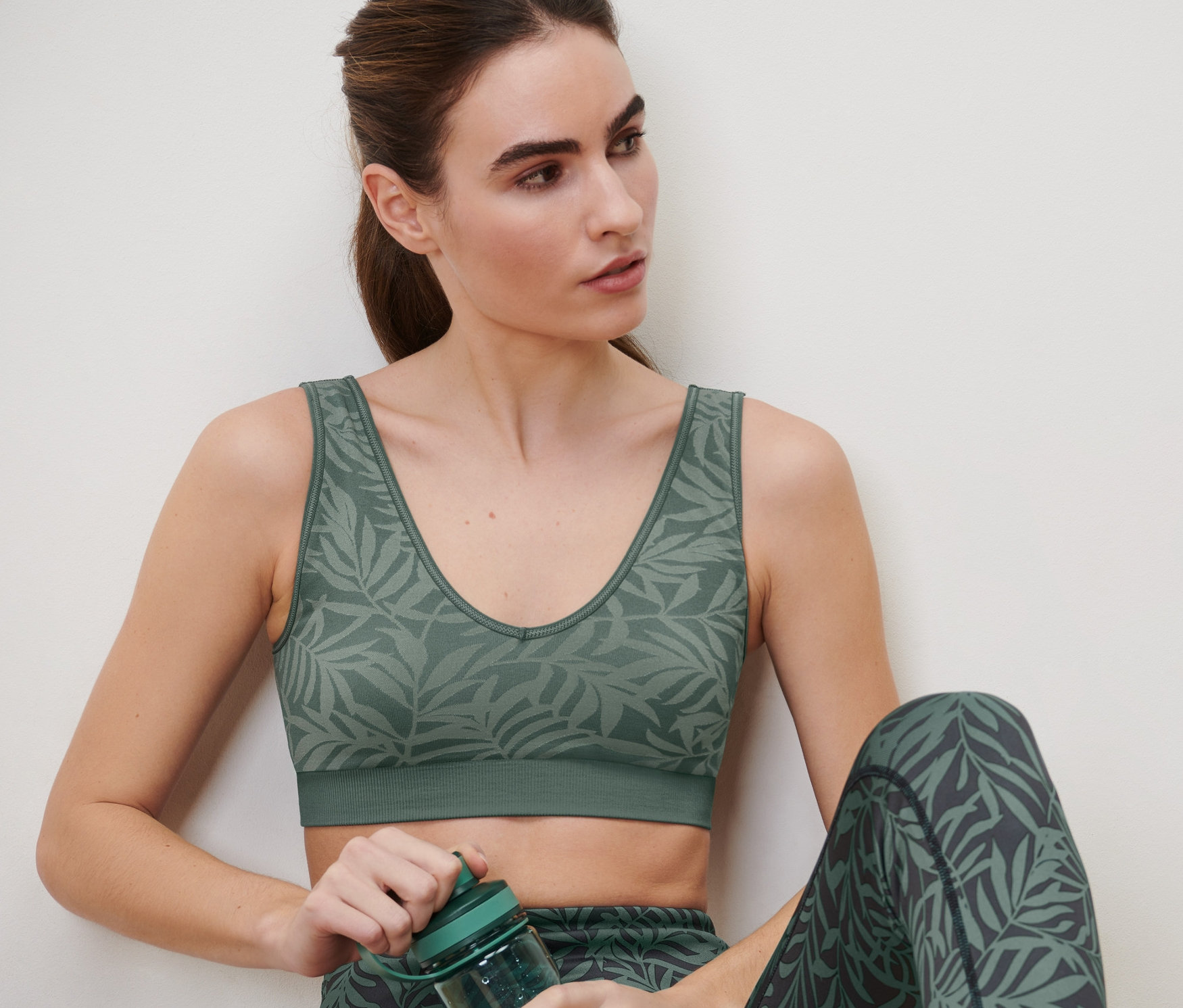 Kobieta siedzi i trzyma zieloną butelkę sportową. Ma na sobie bezszwowy biustonosz sportowy w kolorze szałwiowym i legginsy z nadrukiem liści.