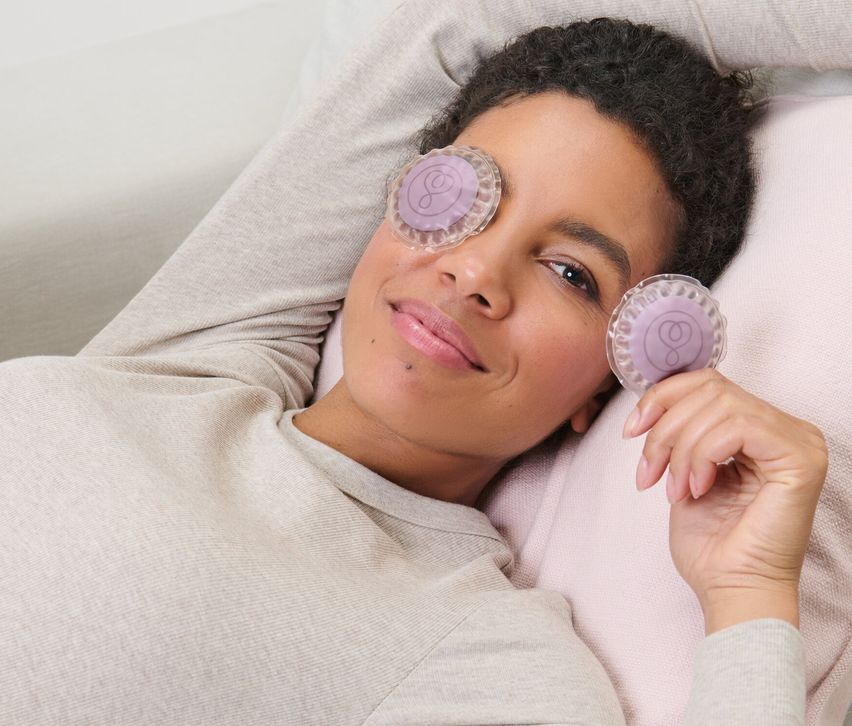 Kobieta leży na kanapie z chłodzącymi okładami na oczy, 2 sztuki, jeden ma na oku, a drugi trzyma w ręku.