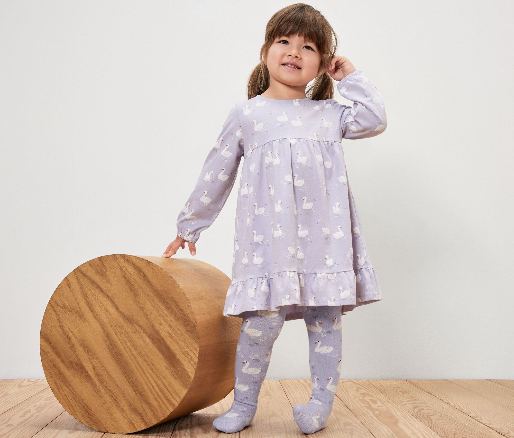 Dziewczynka w sukience z łabędziami stoi obok drewnianego cylindra i ma na sobie Rajstopy dziecięce, 5 par, księżniczka.