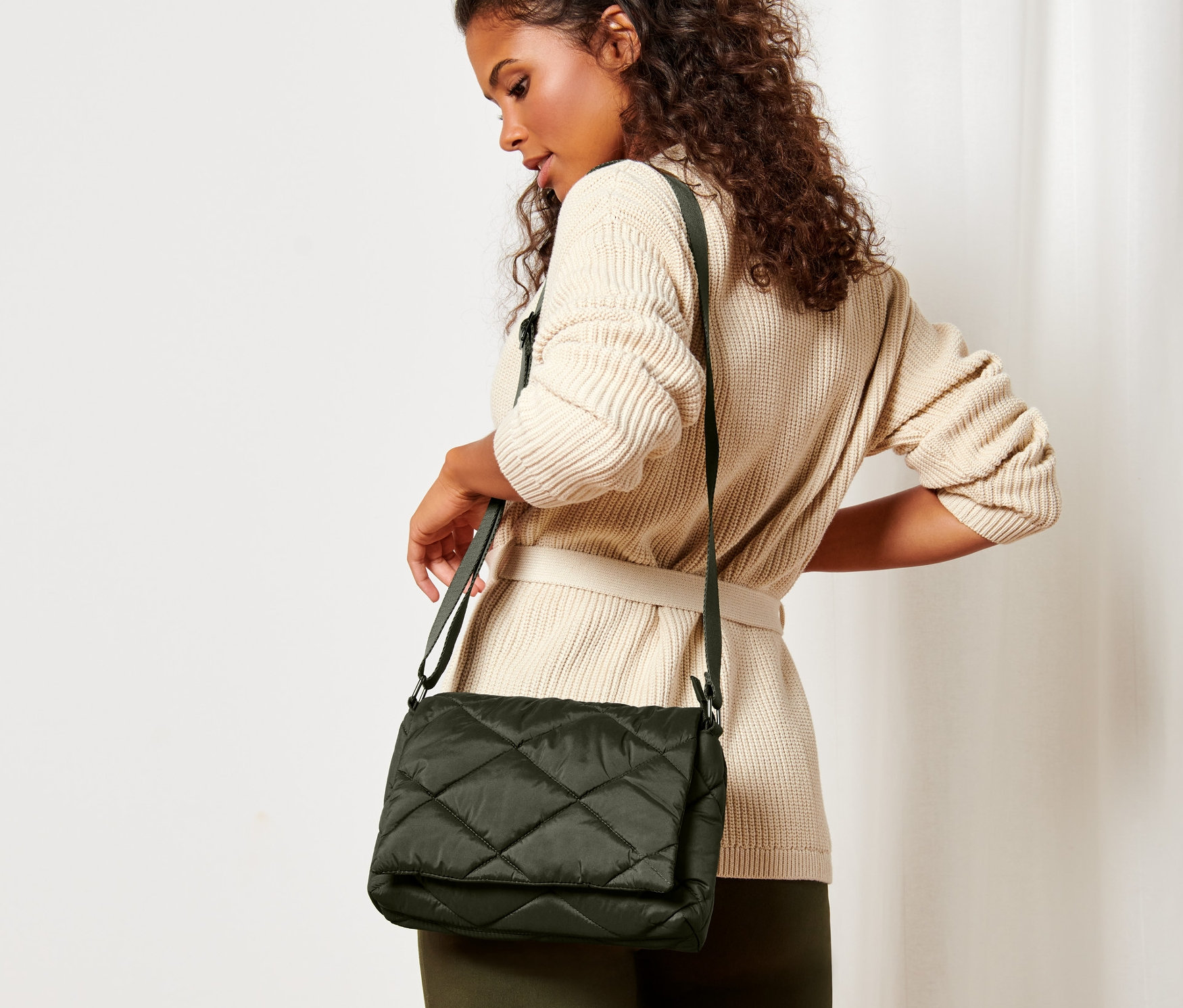 Kobieta o brązowych kręconych włosach w beżowym swetrze z paskiem pozuje z pikowaną torbą na ramię.