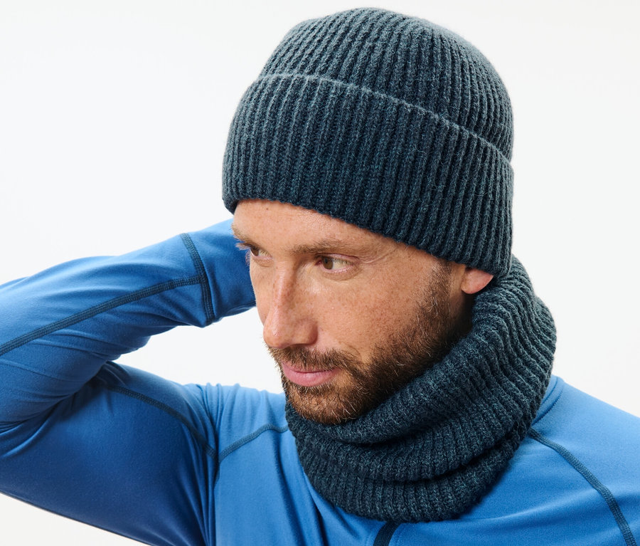 Mężczyzna w niebieskim swetrze i kominie z dzianiny poprawia czapkę.