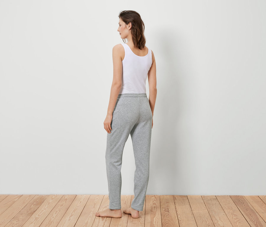 Kobieta stoi tyłem do kamery, ubrana w biały podkoszulek i szare spodnie dresowe.
