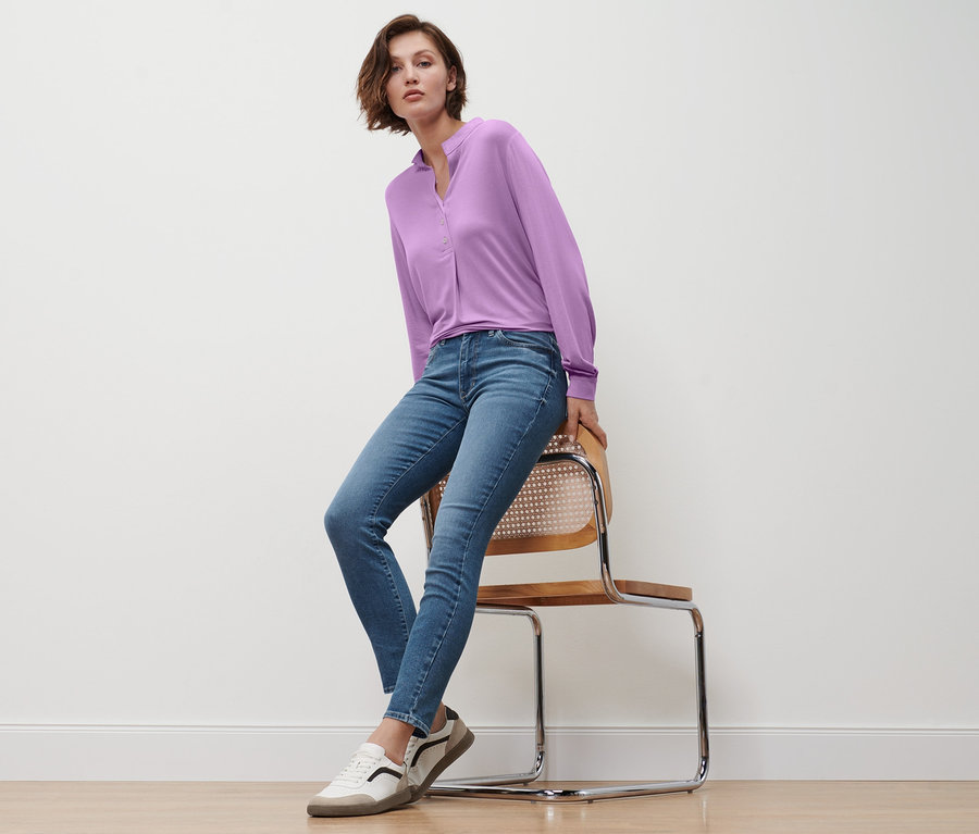 Kobieta siedzi na krześle, ubrana w jeansy slim »Fit Emma«, bluzkę z plisą z guzikami w kolorze liliowym i skórzane sneakersy.