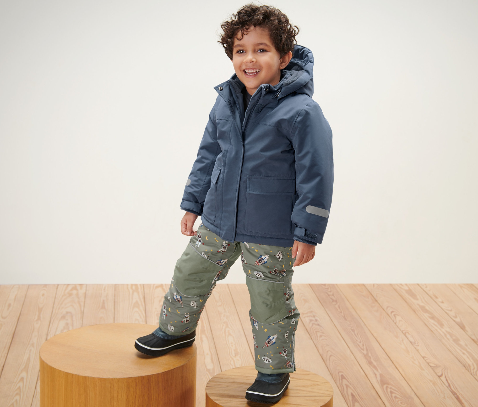 Chłopiec stoi na drewnianym podeście i ma na sobie Dziecięcy dwuczęściowy kombinezon zimowy.