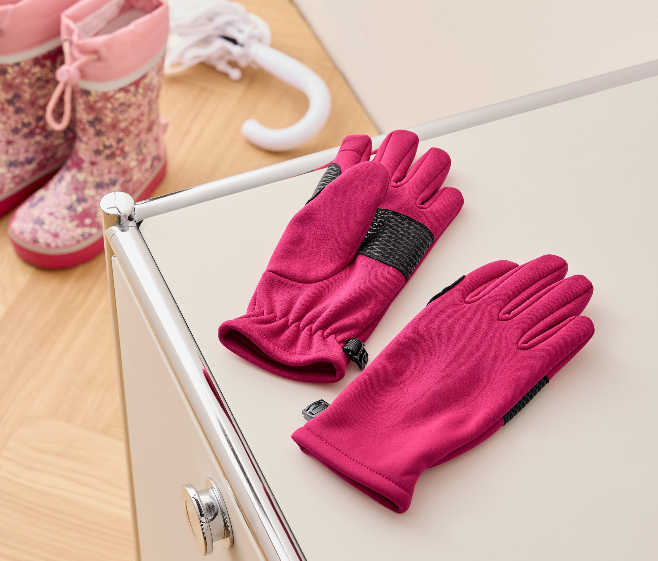 Para różowych rękawic leży na kremowym stoliku obok różowych kaloszy dziecięcych i białego parasola.
