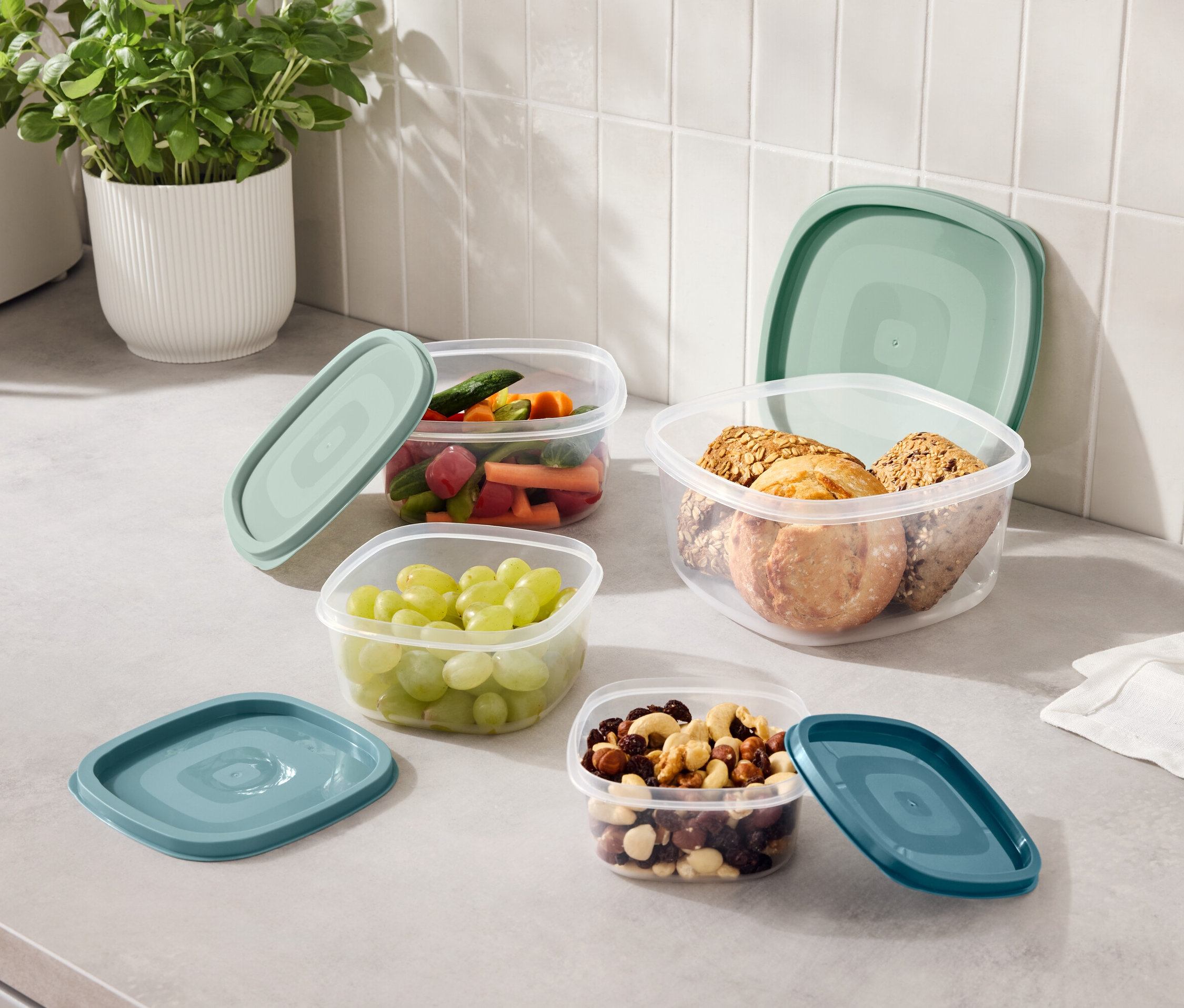 Na blacie kuchennym stoją cztery przezroczyste pojemniki z niebieskozielonymi pokrywkami, wypełnione winogronami, pokrojonymi warzywami, orzechami i ciasteczkami.