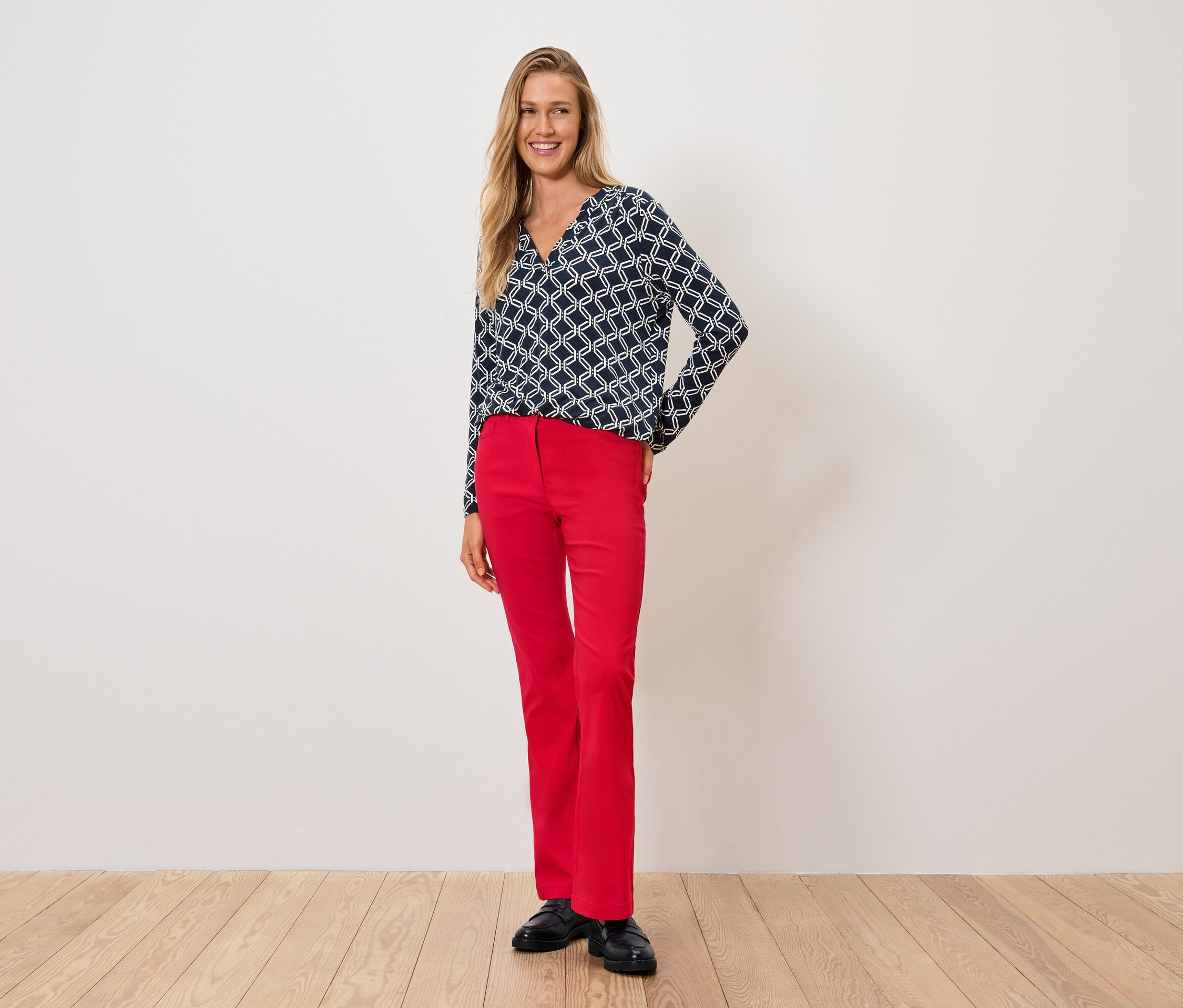 Kobieta w niebiesko-białej bluzce i czerwonych spodniach stoi na drewnianej podłodze.