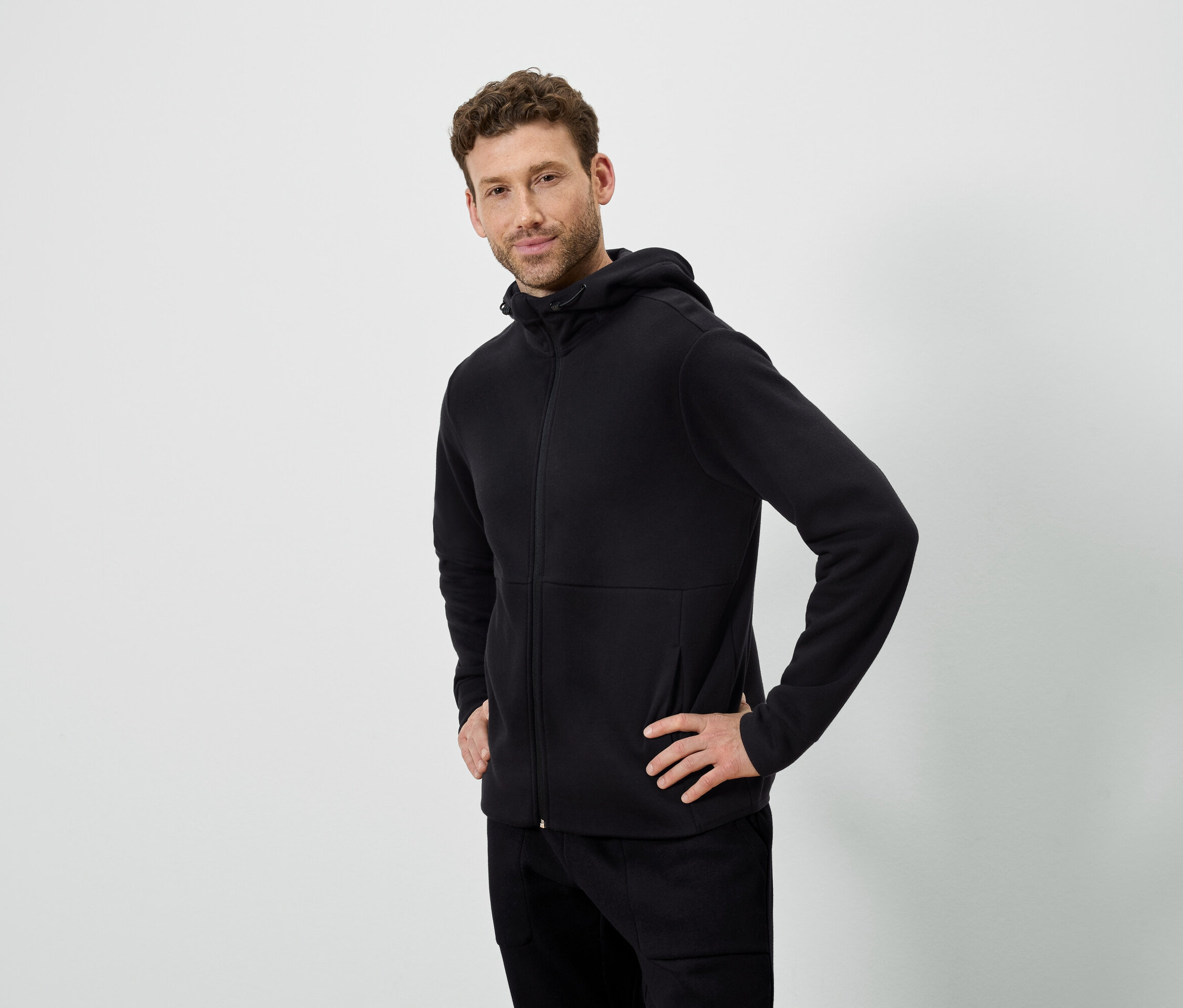Mężczyzna pozuje na białym tle. Ma na sobie czarną bluzę z kapturem i czarne spodnie.