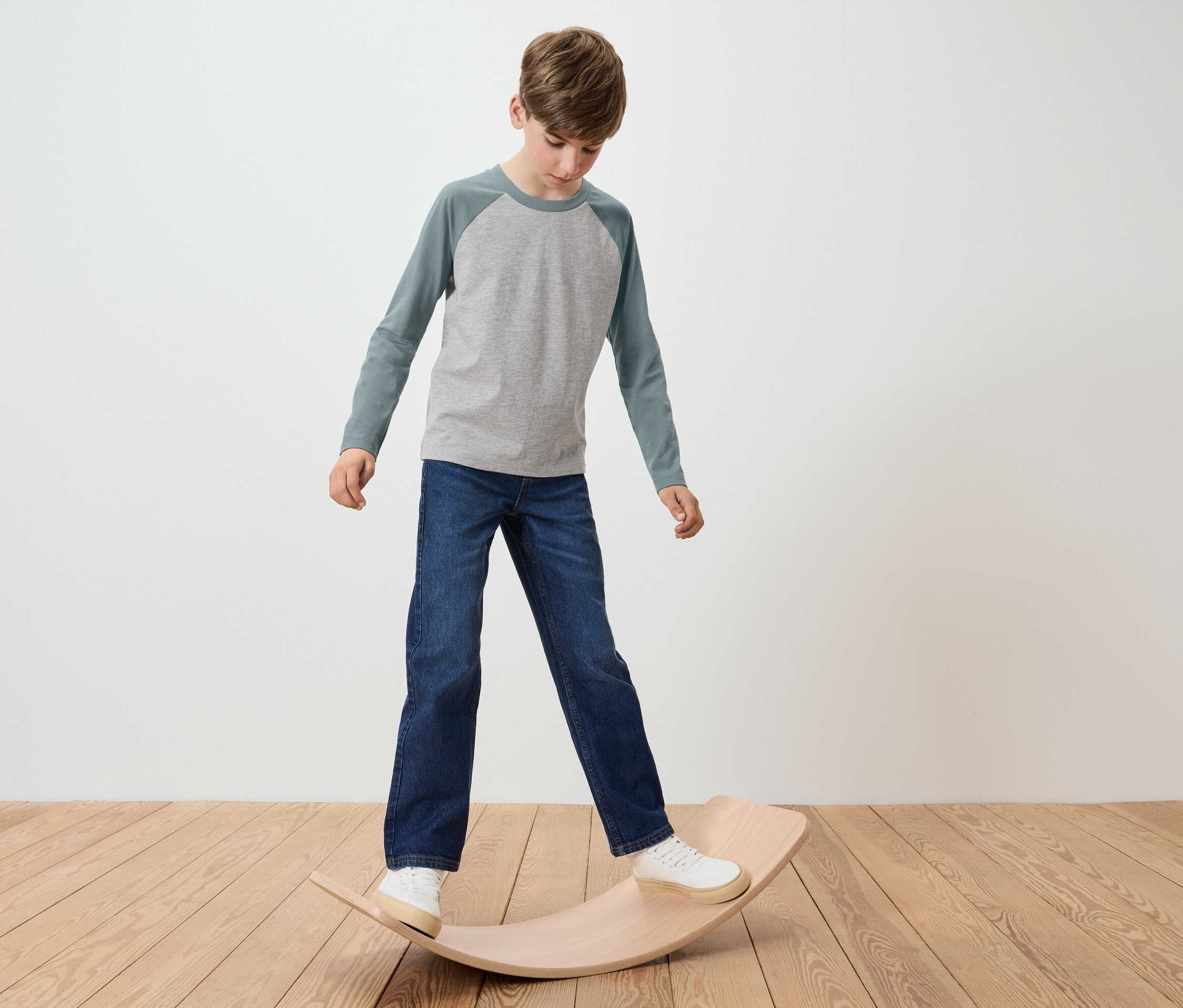 Chłopiec stoi na drewnianej desce do balansowania, ma na sobie szarą koszulkę i dżinsy.