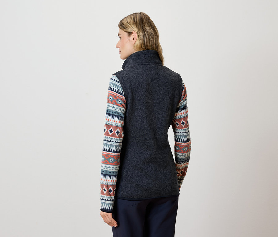 Kobieta tyłem do kamery ma na sobie ciemnoszarą kamizelkę polarową i niebieskie spodnie.