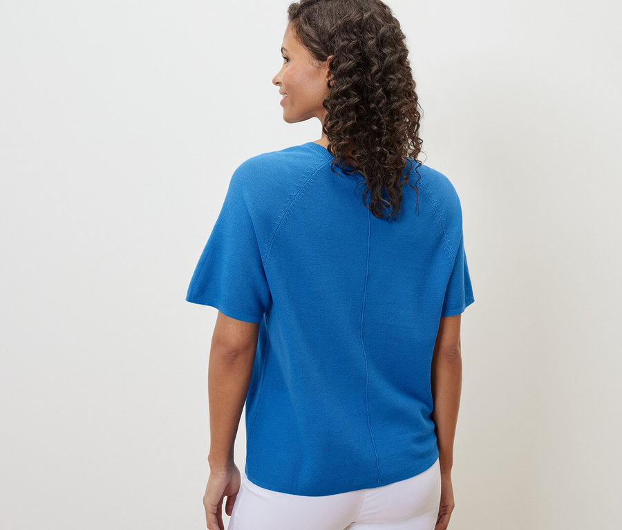 Kobieta widziana od tyłu w kobaltowej bluzce z cienkiej dzianiny i białych spodniach.