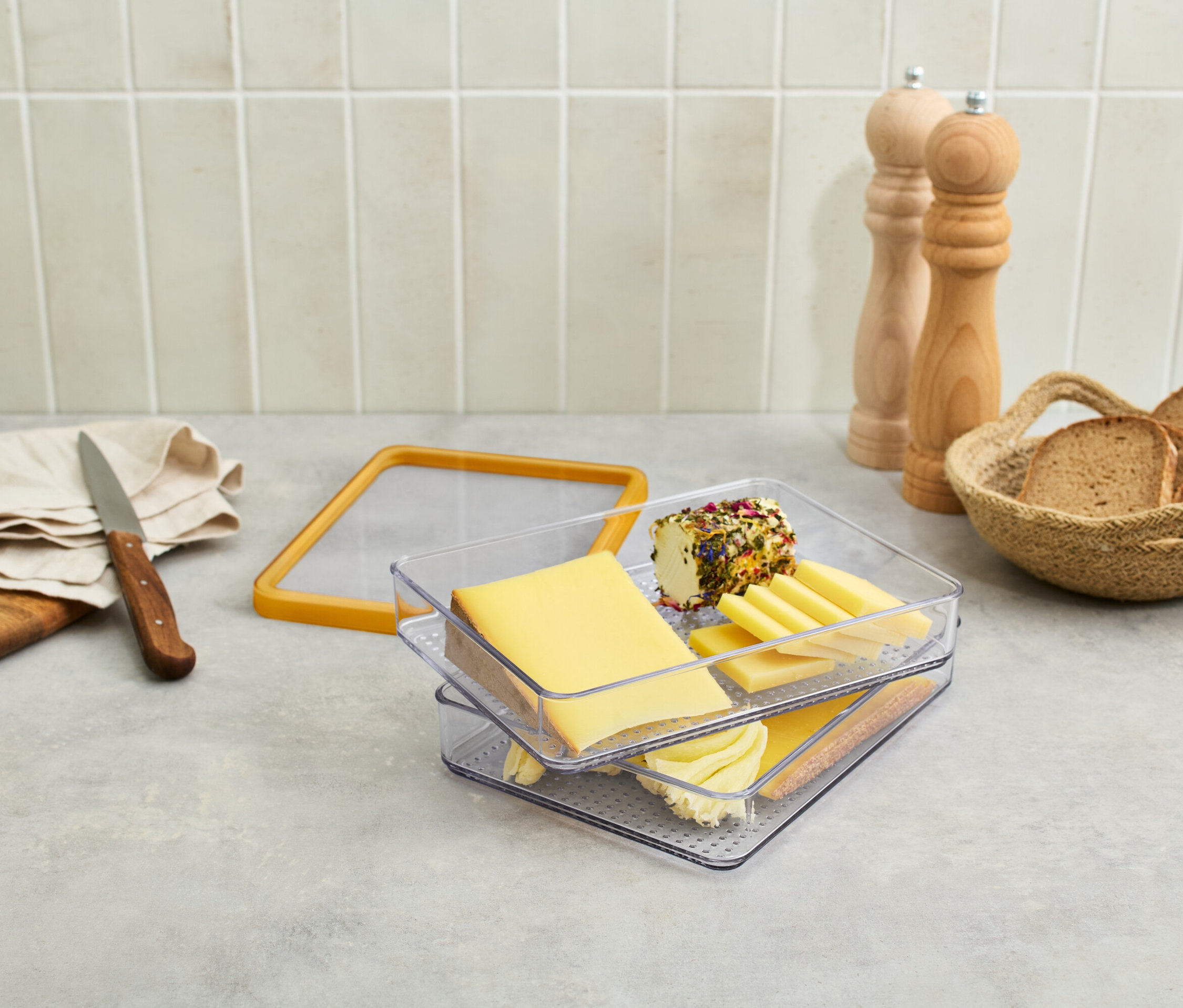 Na stole stoi ser w przezroczystym pojemniku, winogrona, nóż, chleb w koszyku i młynki do przypraw.