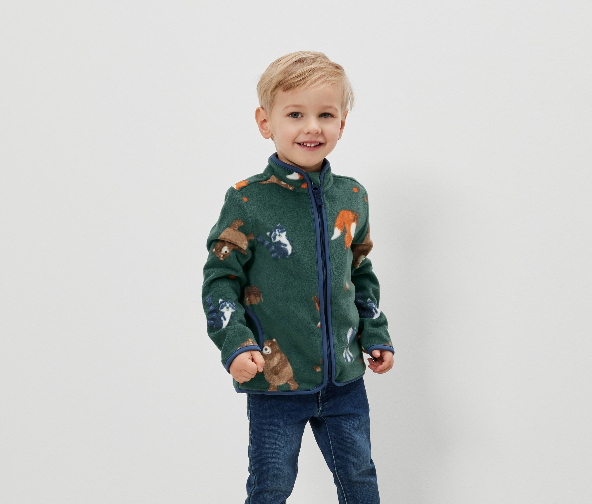 Blondwłosy chłopiec uśmiecha się i pozuje w zielonej bluzie polarowej z nadrukiem zwierzęcym i niebieskimi brzegami.