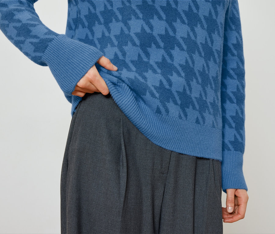Osoba poprawia niebieski sweter w pepitkę i ma na sobie szare spodnie.