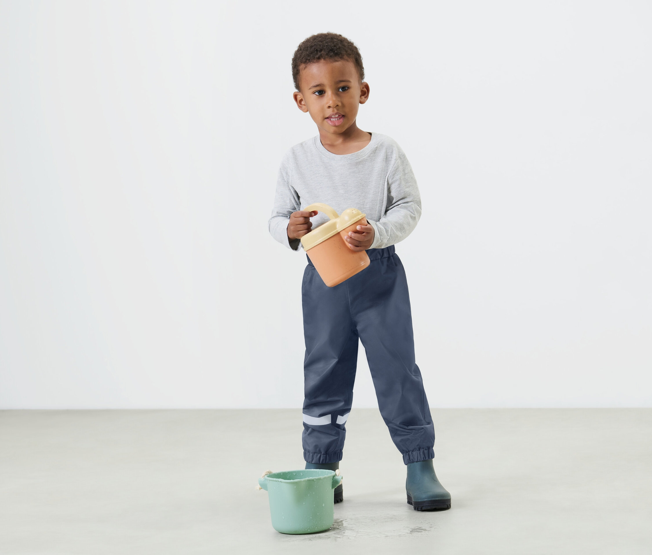 Mały chłopiec trzyma w rękach pomarańczowe wiadro, ubrany w niebieskie spodnie przeciwdeszczowe i kalosze.