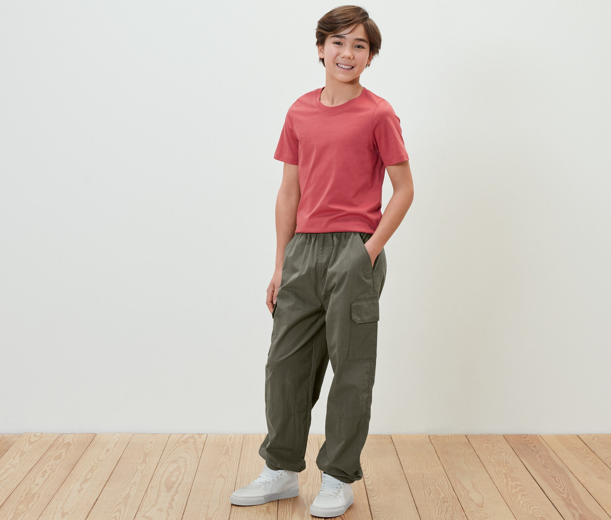 Chłopiec w czerwonym T-shircie i spodniach bojówkach stoi na tle białej ściany.