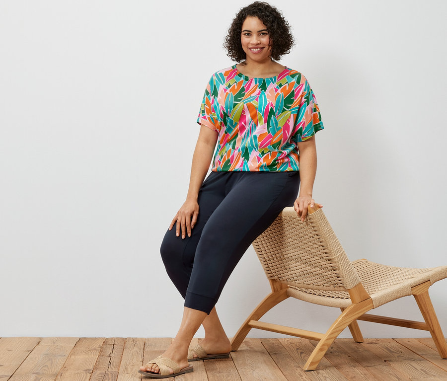 Kobieta siedzi na wiklinowym krześle i ma na sobie granatowe spodnie dżersejowe o długości 3/4 oraz bluzkę z obniżoną linią ramion, ze wzorem na całej powierzchni.
