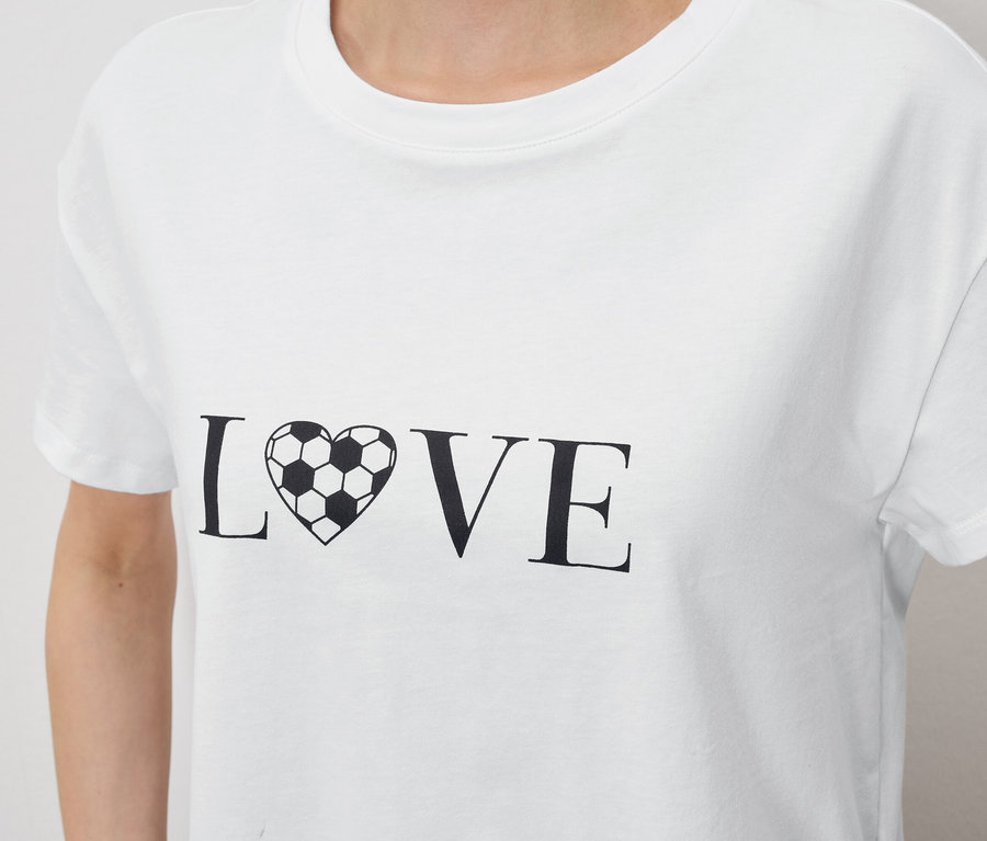 Zbliżenie na osobę ubraną w białą koszulkę z napisem "LOVE", gdzie "O" zastąpione jest sercem w kształcie piłki nożnej.
