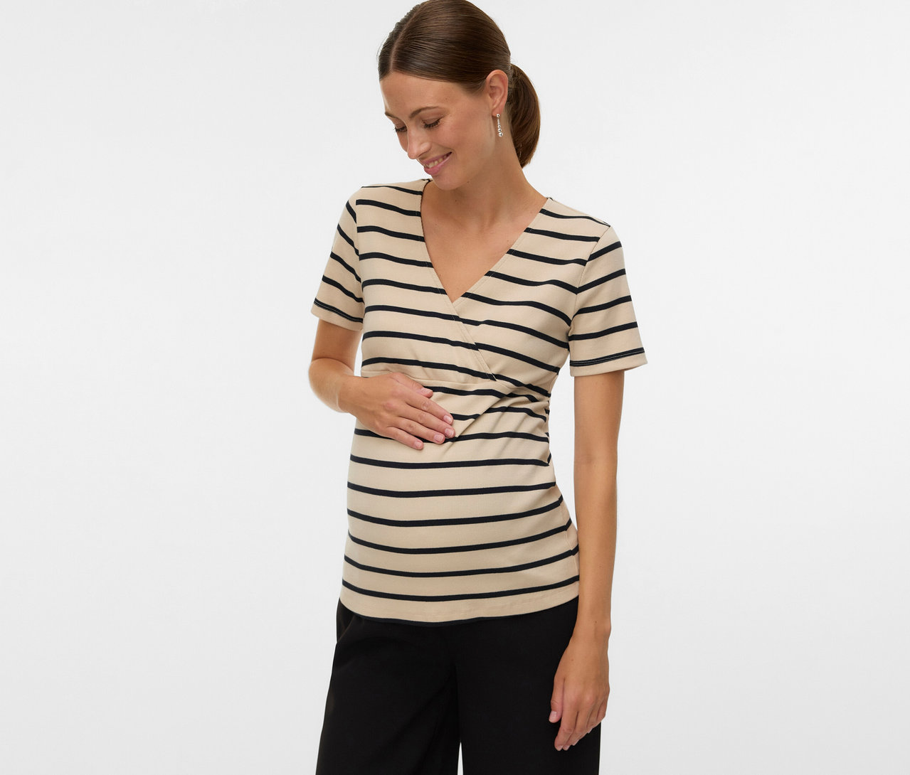 Kobieta z końskim ogonem ma na sobie koszulkę ciążową MAMALICIOUS „MLLANE TESS” w paski i czarne spodnie, dotykając brzucha.