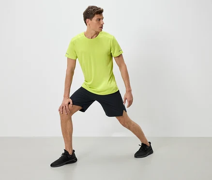 Mężczyzna w odzieży sportowej rozciąga się. Ma na sobie neonowo-zielony t-shirt, czarne szorty i czarne buty sportowe.