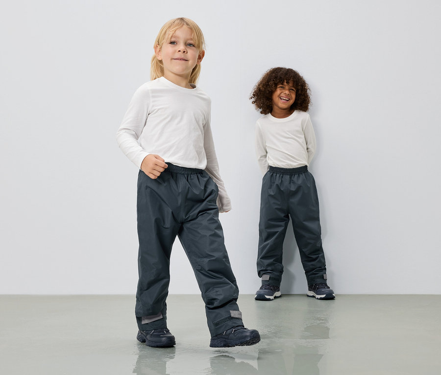 Dwoje dzieci ma na sobie długie, szare spodnie przeciwdeszczowe i białe bluzki z długim rękawem na tle białej ściany.