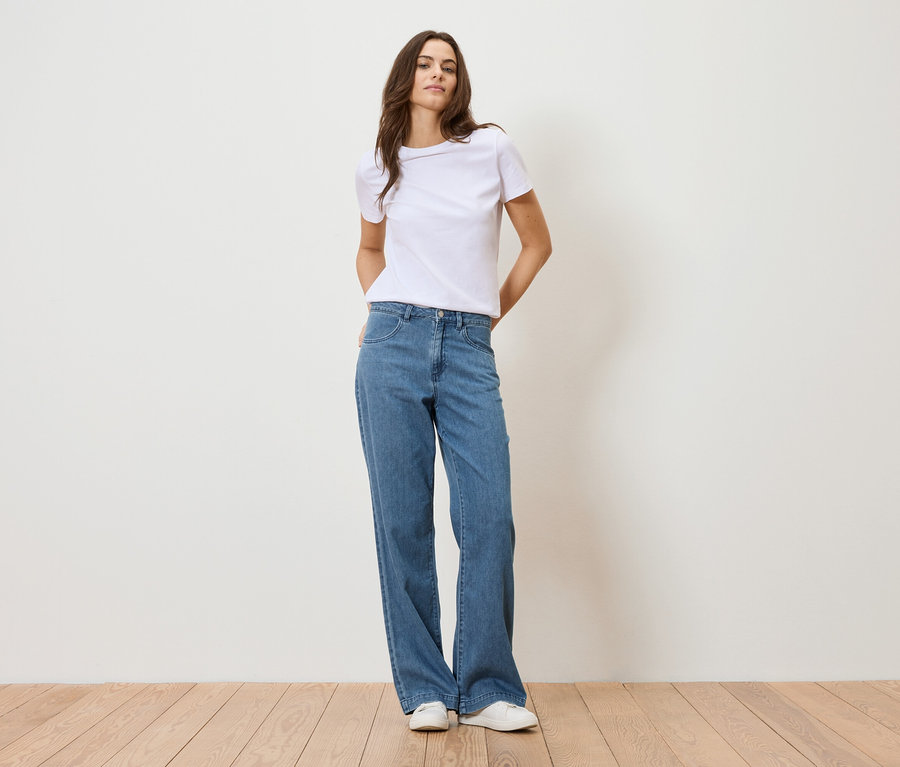 Kobieta stoi na tle białej ściany, ubrana w białą koszulkę i niebieskie dżinsy.