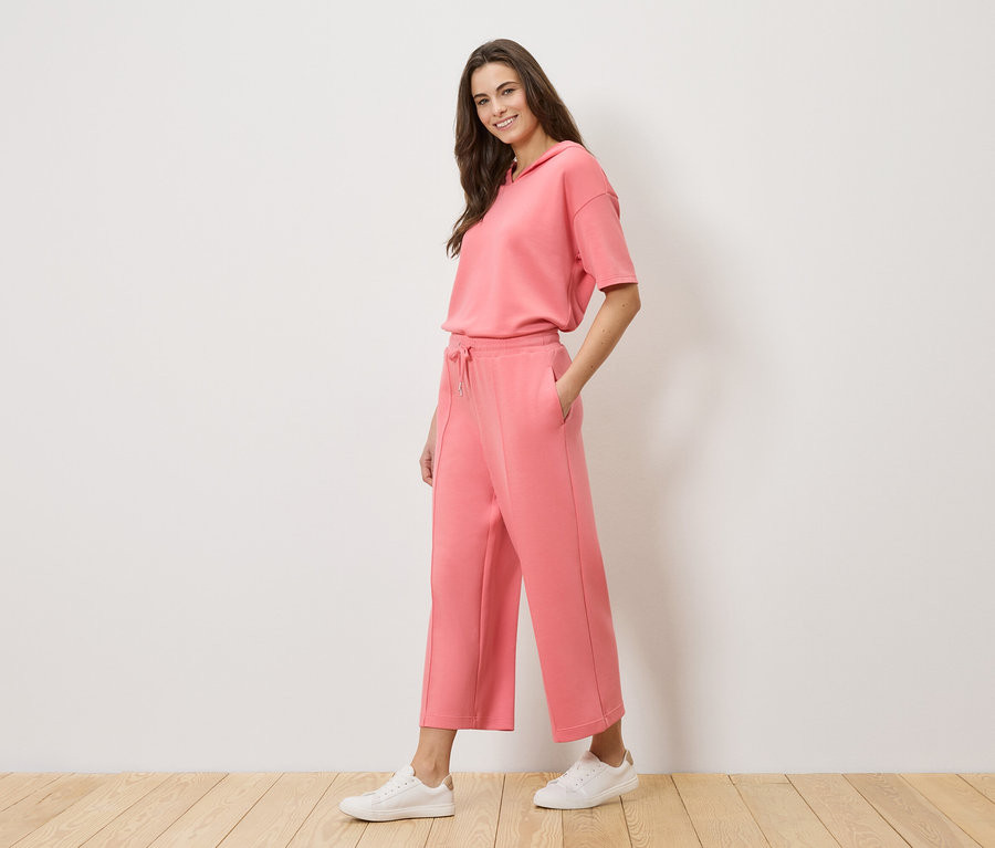 Kobieta ma na sobie koralowy dres z bluzą z kapturem i kulotami oraz białe tenisówki.