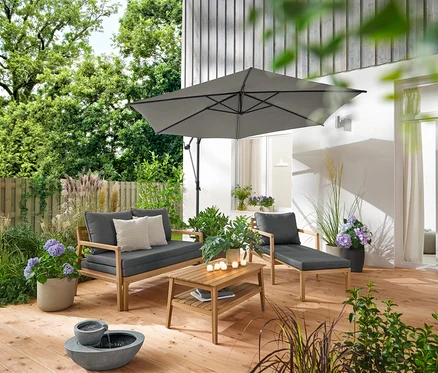 Na drewnianym tarasie stoją rozsuwany fotel ogrodowy, rozsuwana sofa ogrodowa, ogrodowy stolik kawowy (ok. 80 x 50 cm) i solarna fontanna ogrodowa. Parasol Leco w kolorze antracytowym zapewnia cień. 2 poduszki ogrodowe o strukturze lnu i 6 akumulatorowych lampek LED do użytku na zewnątrz zdobią otoczenie.