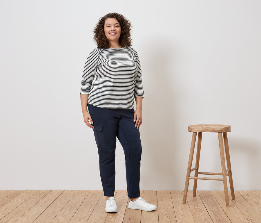 Kobieta z kręconymi włosami stoi w bluzce w paski, niebieskich spodniach i białych tenisówkach obok stołka.