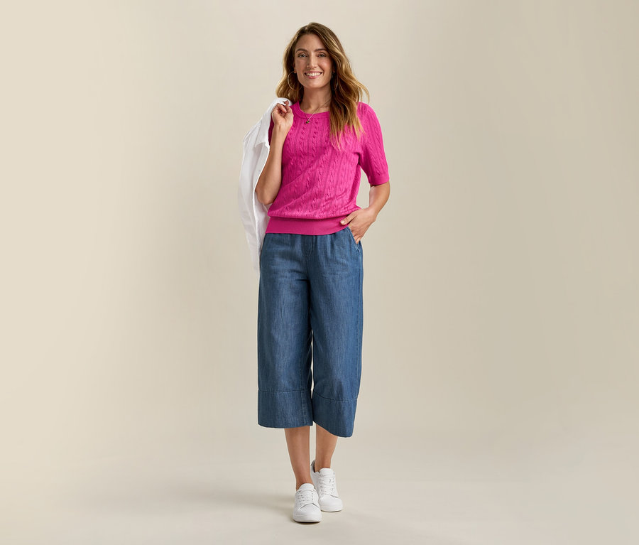 Uśmiechnięta kobieta ma na sobie dżinsową kurtkę i różowy, cienki sweter z warkoczem. Nosi niebieskie culottes i białe trampki.