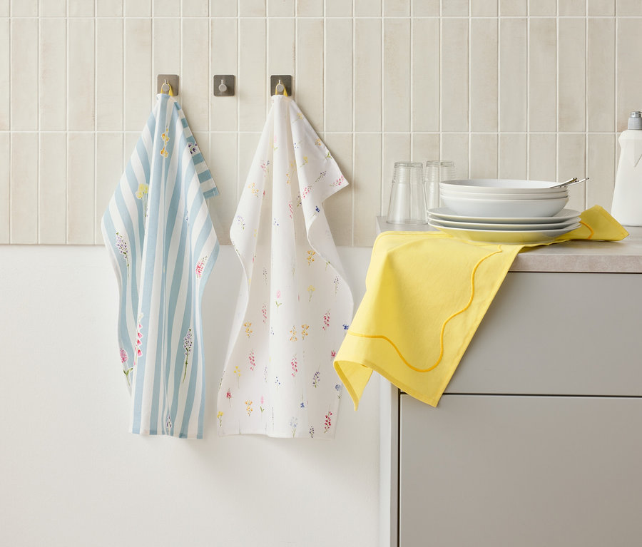 Dwa ręczniki kuchenne wiszą na haczykach na ścianie nad blatem kuchennym, na którym znajdują się naczynia, szklanki, butelka płynu do mycia naczyń i żółta ściereczka.