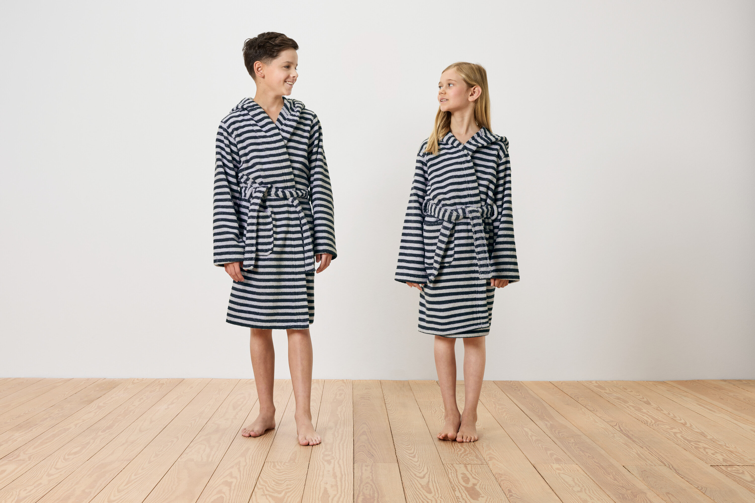 Chłopiec i dziewczynka stoją boso na drewnianej podłodze, ubrani w pasiasty szlafrok z kapturem.