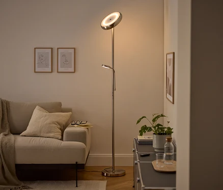 Nowoczesna srebrna lampa podłogowa z głównym światłem skierowanym w górę i regulowaną lampką do czytania oświetla przytulny salon. Widoczne są beżowa sofa z kocem i poduszką, dwa oprawione obrazy line art oraz ciemna szafka z rośliną i butelką wody.