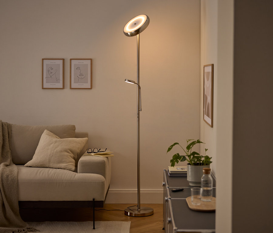 Nowoczesna srebrna lampa podłogowa z głównym światłem skierowanym w górę i regulowaną lampką do czytania oświetla przytulny salon. Widoczne są beżowa sofa z kocem i poduszką, dwa oprawione obrazy line art oraz ciemna szafka z rośliną i butelką wody.