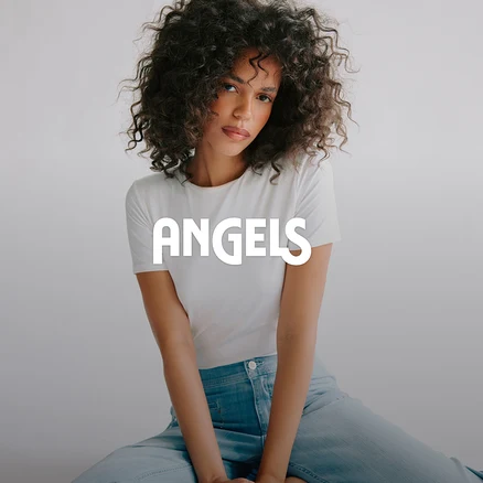 Kobieta z kręconymi włosami w białym T-shircie i jasnoniebieskich dżinsach siedzi i patrzy w kamerę. Na zdjęciu widnieje napis ANGELS.
