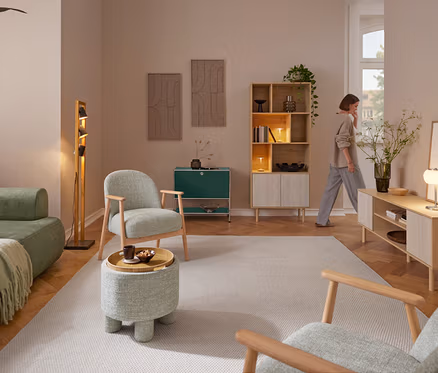 Kobieta przechodzi przez salon urządzony zieloną sofą, dwoma fotelami na drewnianej ramie i pufą do siedzenia z tacą. Obok sofy stoi lampa stojąca Trio „Bell”. Przy tylnej ścianie znajduje się metalowa szafka typu sideboard „CN3” i regał na książki w imitacji trawertynu z dwiema lampami stołowymi LED Trio „Madison”. Po prawej stronie stoi szafka typu lowboard w imitacji trawertynu z lampą stołową LED Trio „Madison” i wazonem z kwiatami.