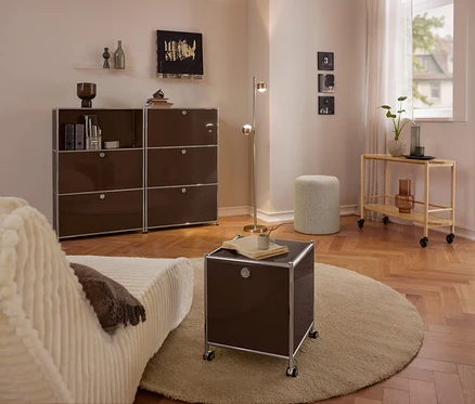 Nowoczesny salon z beżową sofą, dużą brązową metalową komodą »CN3« z 3 klapami, małą brązową metalową komodą »CN3« na kółkach z 2 klapami na okrągłym dywanie, pufem Bouclé i drewnianym wózkiem do serwowania.