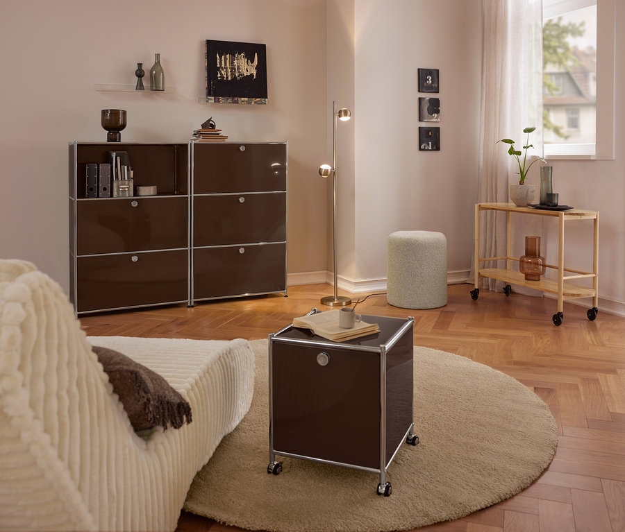 Nowoczesny salon z beżową sofą, dużą brązową metalową komodą »CN3« z 3 klapami, małą brązową metalową komodą »CN3« na kółkach z 2 klapami na okrągłym dywanie, pufem Bouclé i drewnianym wózkiem do serwowania.