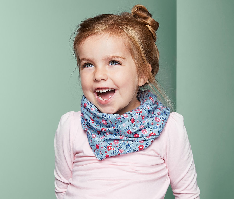 Uśmiechnięta dziewczynka siedzi i ma na sobie niebieską chustkę wielofunkcyjną z kwiatowym wzorem.