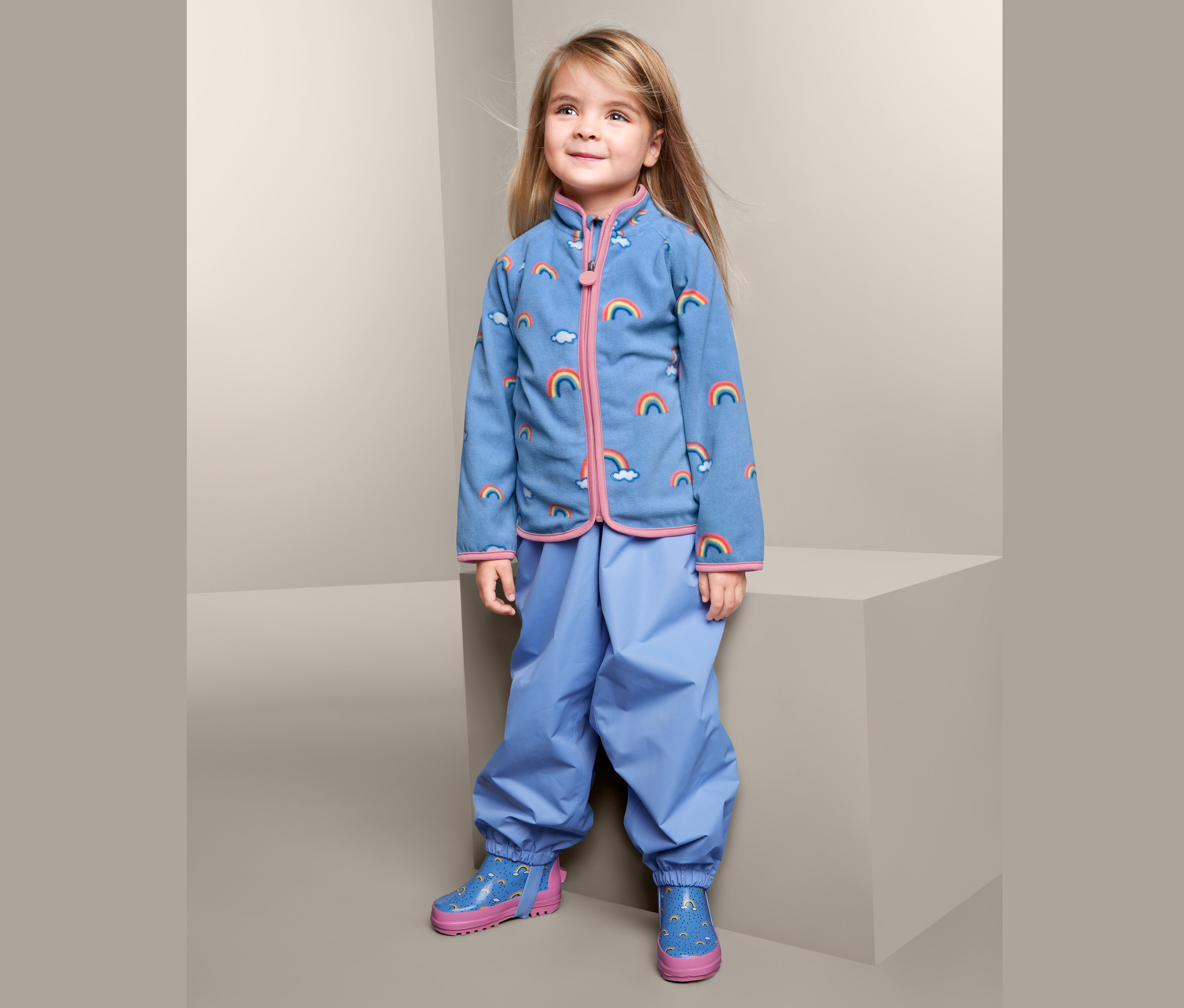 Dziewczynka ma na sobie rozpinaną bluzę z mikropolaru w tęczowy wzór, niebieskie spodnie i kalosze w tęczowy wzór.