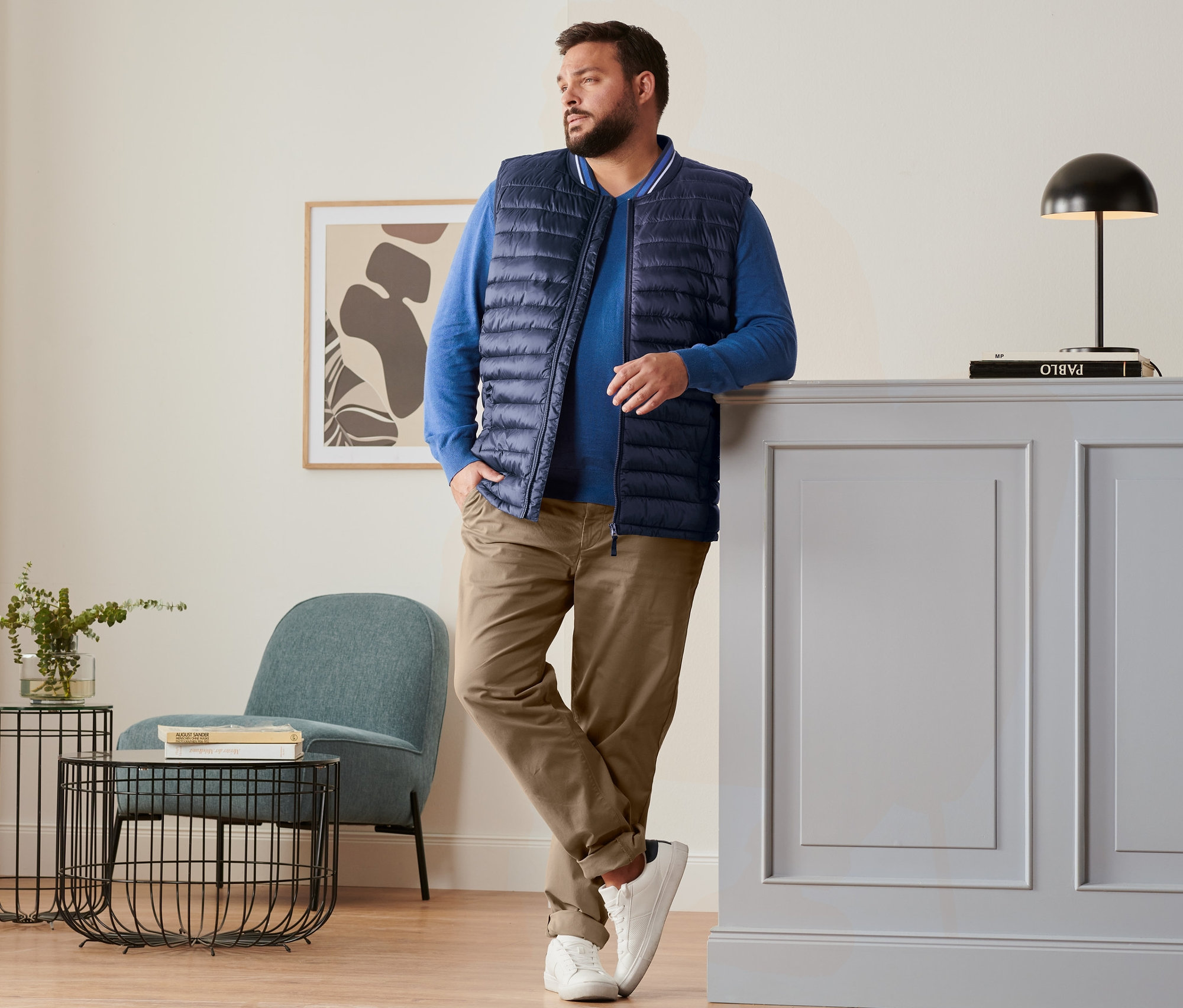 Mężczyzna opiera się o komodę, ubrany w niebieską kamizelkę pikowaną, niebieski sweter lniany, karmelowe spodnie chino i buty typu sneaker.